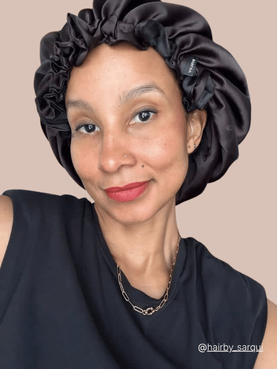 A close-up portrait of a woman wearing a black satin BUNLUV bonnet , paired with a black sleeveless top and a delicate gold chain necklace, set against a solid beige background.