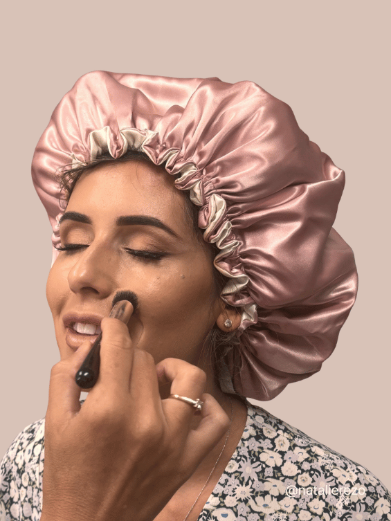 A woman wearing a BUNLUV satin sleep bonnet in dusty pink, applying makeup with a brush. The background is a solid warm beige color.