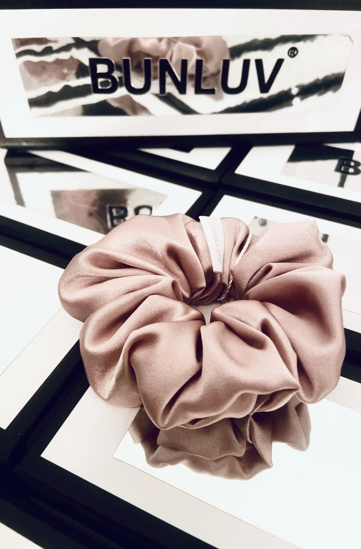 A dusty rose bunluv satin hair scrunchie displayed on a black and white geometric surface, with a branded BUNLUV box partially visible in the background.