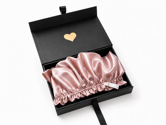 Pink satin bonnet in a black box with a gold heart emblem on a white background
