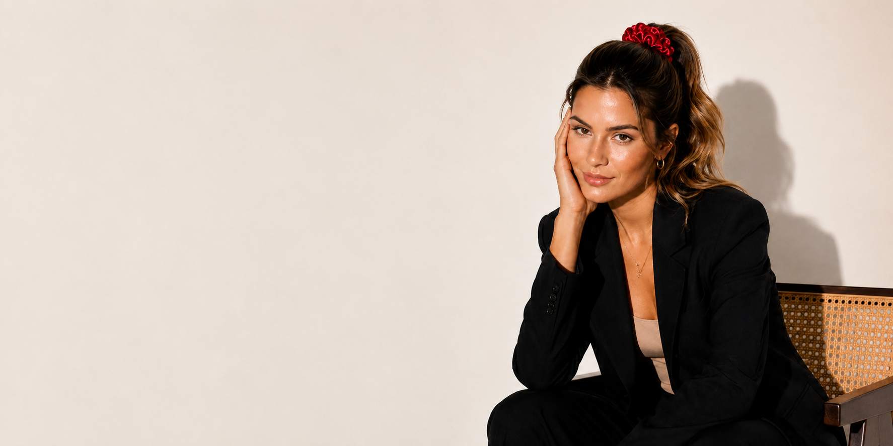 A woman with her hair in a high ponytail sits on a woven chair against a plain off-white wall, wearing a black blazer over a light beige top and a red satin bunluv scrunchie 